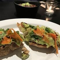 Two pieces of hard bread with green and orange foodstuff on them on a white plate with a bowl and glass on the table in the background