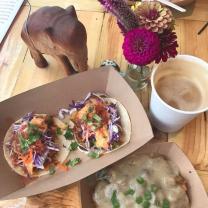 A top down view of a meal - a brown holder with what looks like two taco-like things, and brown drink in a white cup, something in a brown holder at the bottom, white potatoes with something green sprinkled on top and a red flower in a vase and an elephant statue