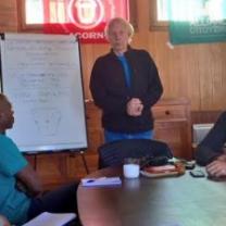 Gray haired thin man standing by a white paper flip chart in front of a group of people at a table