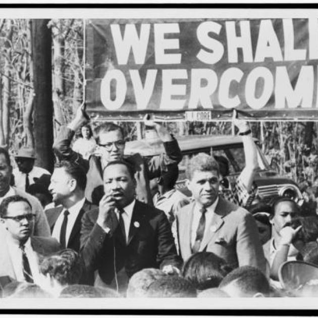 Black and white photo of Martin Luther King Jr. and others marching under sign that says We Shall Overcome