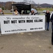 People outside holding a long white banner with black letters saying We remember the victims...But not with more killing and www.abolition.org and a phone number