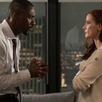 On the left a black man facing right in a white shirt and tie looking at a woman his hands in front of him with fingers clasped like he's praying and the white woman with long brown hair to the left is looking up at him with arms folded wearing a pick shirt. They are standing in front of a window looking over a nighttime city scene