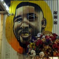 Painting of a black man's face against a yellow and orange circle on a white sided building with a bouquet of flowers in the foreground