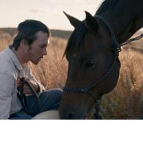 Young man with brown hair looking sideways and a big brown horse leaning its head toward his lap