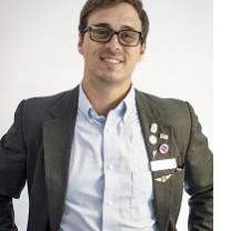 Young white man with dark rimmed glasses and a suit with lots of political buttons and a big smile