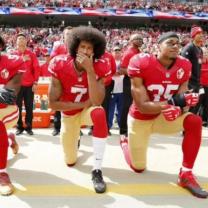 African American football players with red jerseys and tan pants kneeling on one knee in a stadium full of people