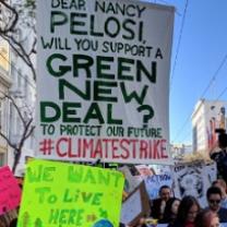 Outdoor rally with signs, one says We Want to Live Here, one says Dear Nancy Pelosi will you support a Green New Deal