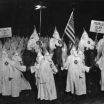 People in white robes with pointy top hats in a circle outside at night with a flag