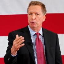 White man middle-aged in dark suit with red tie and white shirt talking and holding up his hand by his chest in front of a red and white striped background