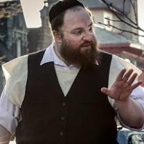 Large white man with jewish hat and wire rimmed glasses, a longish brown beard wearing a white collared button down shirt and black button vest his hand gesturing as he talks to someone to his right