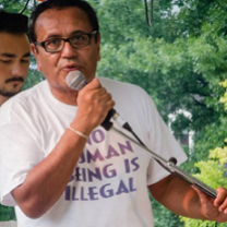 Latino man in T-shirt that says No Human Being is Illegal, talking into a mic