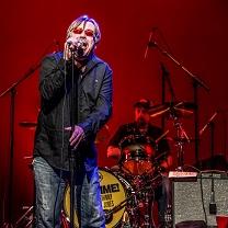 Middle aged white man with red sunglasses on singing with wide mouth open into a standing mic in front of a guy playing drums