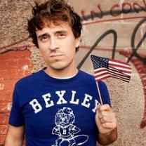 Young brown haired man wearing a blue t-shirt saying Bexley and waving a tiny flag in front of some kind of wall with words on it