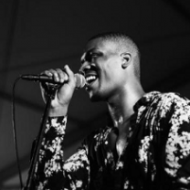 Black and white photo of bald black man smiling and singing into a mic with a flowered shirt on