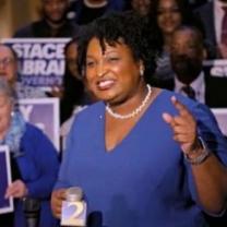 Older black woman with short curly black hair, white pearl necklace, blue dress, standing and smiling while gesturing in front of lots of people holding signs that say Stacey Abrams